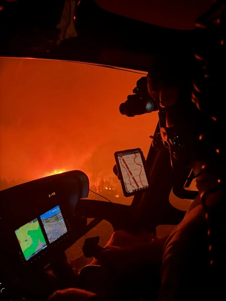 View from a helicopter cockpit toward a wildfire glowing beyond the windshield